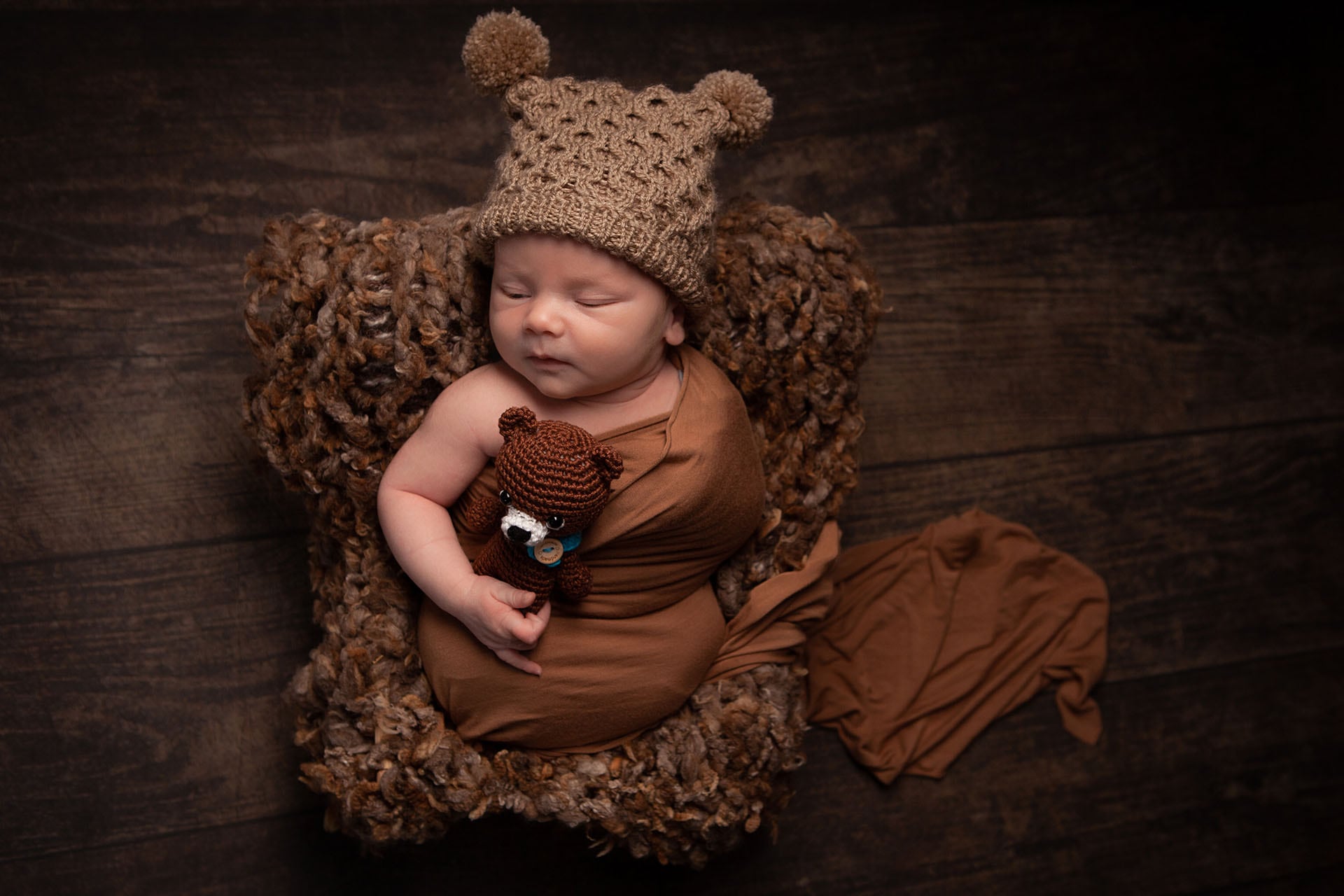 A newborn baby sleeping in a brown blanket with a teddy bear.