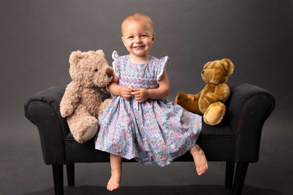 smiling little girl in a pretty dress on a stool with a teddy bear on either side