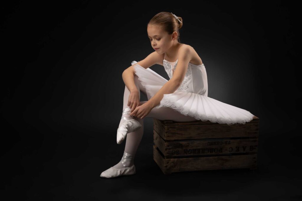 ballerina in a white tutu sitting on a box on a white background sorting the ribbons on her shoes