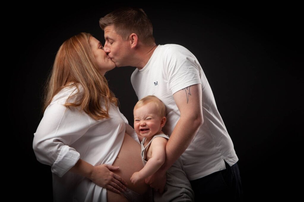 A pregnant woman kisses her partner in front of a black background.