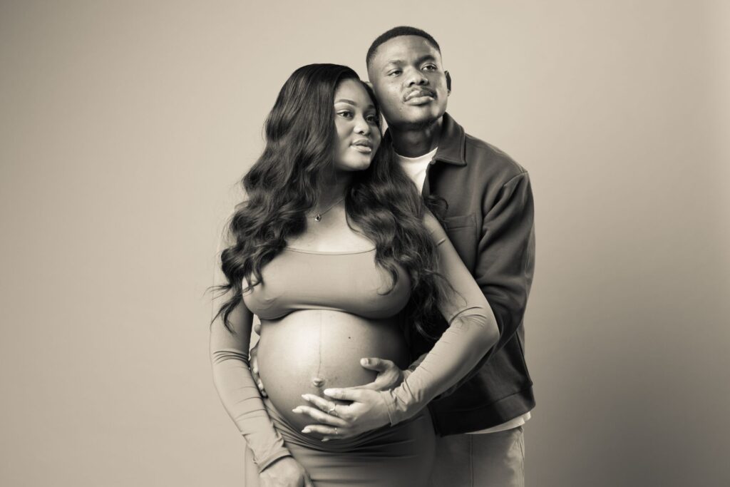 A pregnant woman and a man stand together, the man's arms around her. She cradles her belly. Both look away, appearing thoughtful. Sepia-toned image.