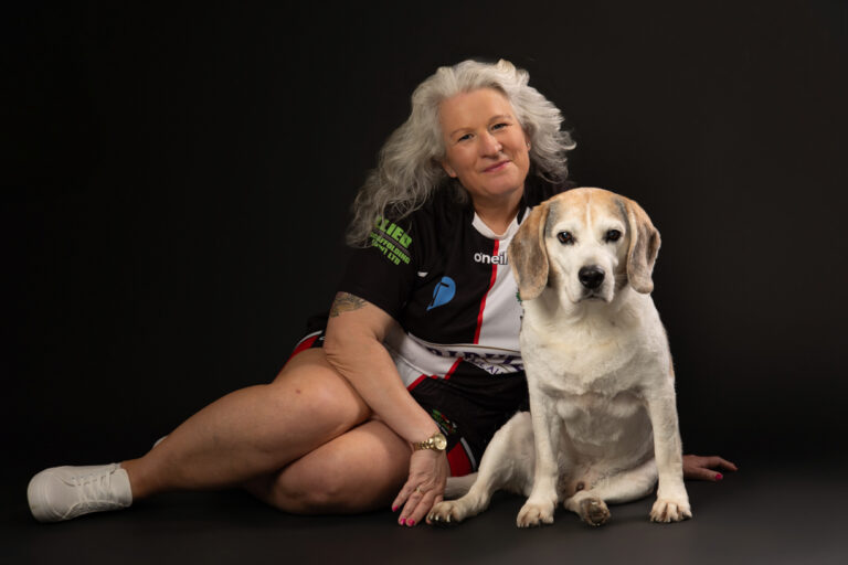 A woman with long grey hair sits on the floor next to a large white and tan dog, both facing forwards against a plain black background.