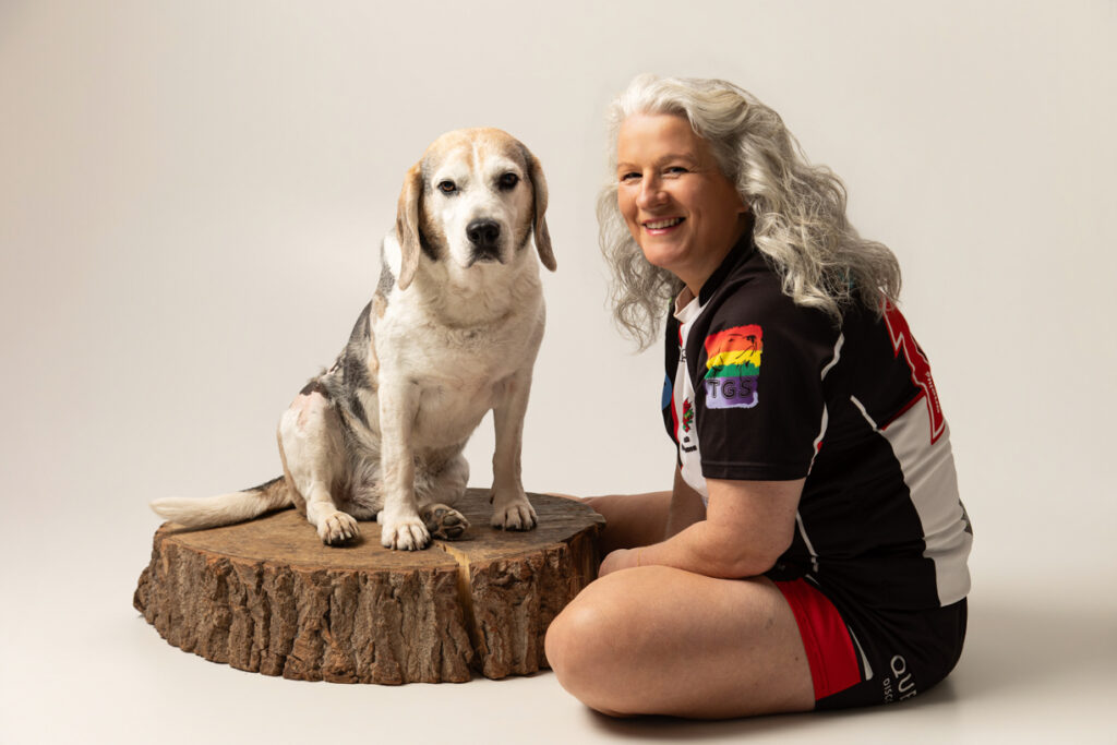 A woman with grey hair sits next to a beagle on a tree stump, both facing the camera. The woman wears a sports jersey with rainbow patches and smiles.