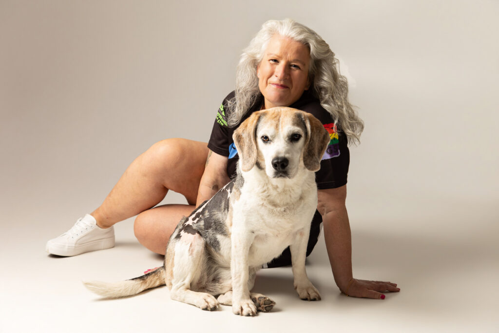 A woman with long grey hair sits on the floor, wearing a dark shirt and white trainers, with a white-and-brown dog sitting in front of her against a plain background.