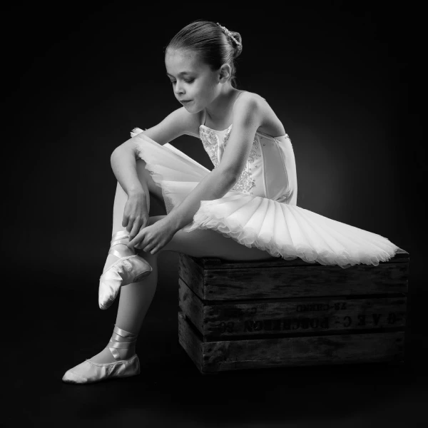 A young ballerina in a tutu sits on a wooden crate, adjusting her pointe shoe, against a dark background.