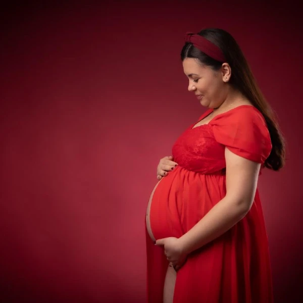A pregnant woman in a red dress stands in profile against a matching red background, gently holding her exposed bump and looking down.