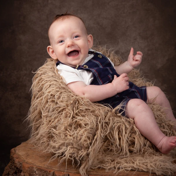 Smiling baby wearing a white shirt and blue checked dungarees sits on a furry chair placed on a tree stump against a brown studio backdrop.