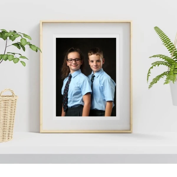 Framed photo of two children in school uniforms, standing side by side and smiling, displayed on a white shelf with potted plants nearby.