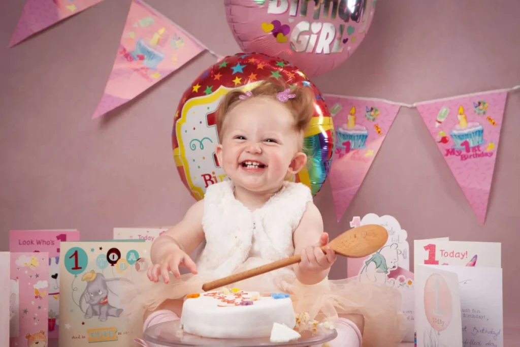 Smiling baby in a white dress holds a wooden spoon and sits in front of a small birthday cake, with balloons, cards, and birthday decorations in the background.