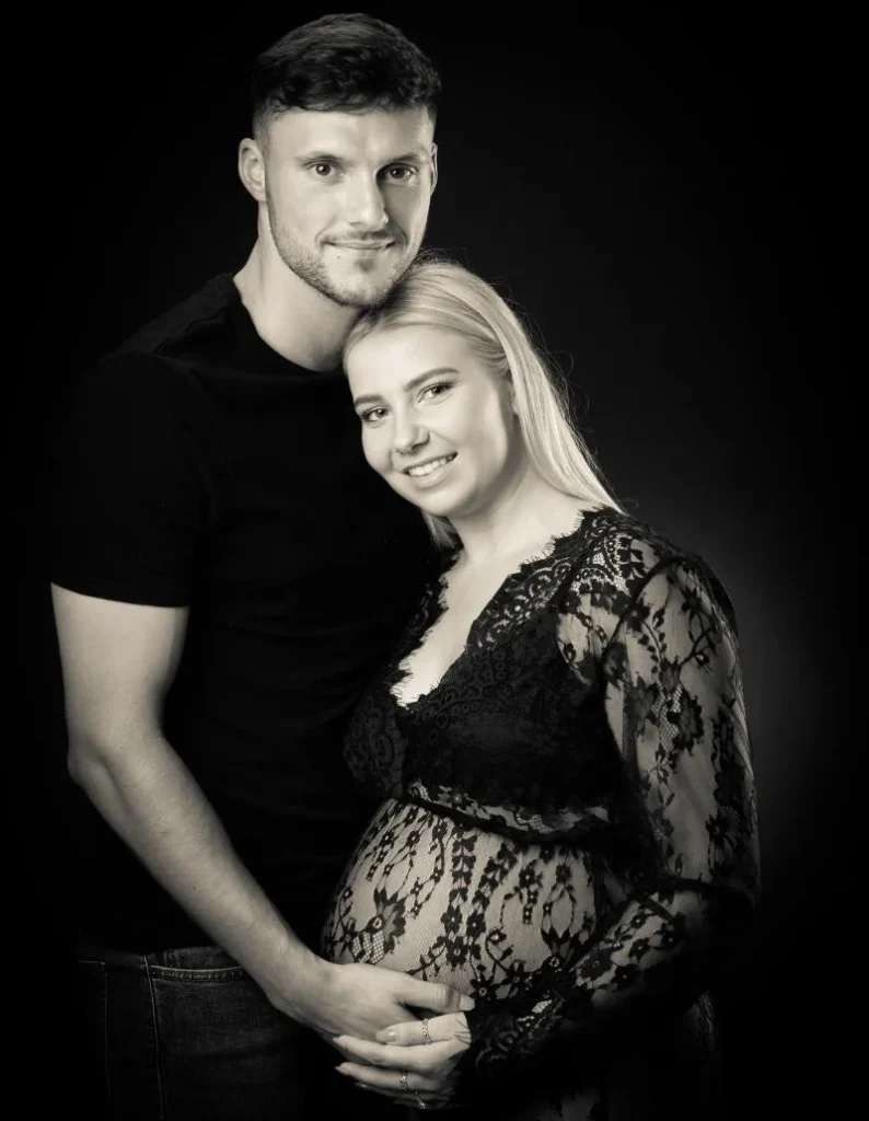 A man stands behind a pregnant woman wearing a lace dress. Both are smiling and holding her baby bump, posing in front of a plain dark background.