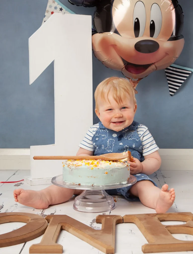 A smiling baby sits in front of a blue iced cake and large white number one, with a Mickey Mouse balloon in the background and the word “ONE” spelt out in wooden letters on the floor.