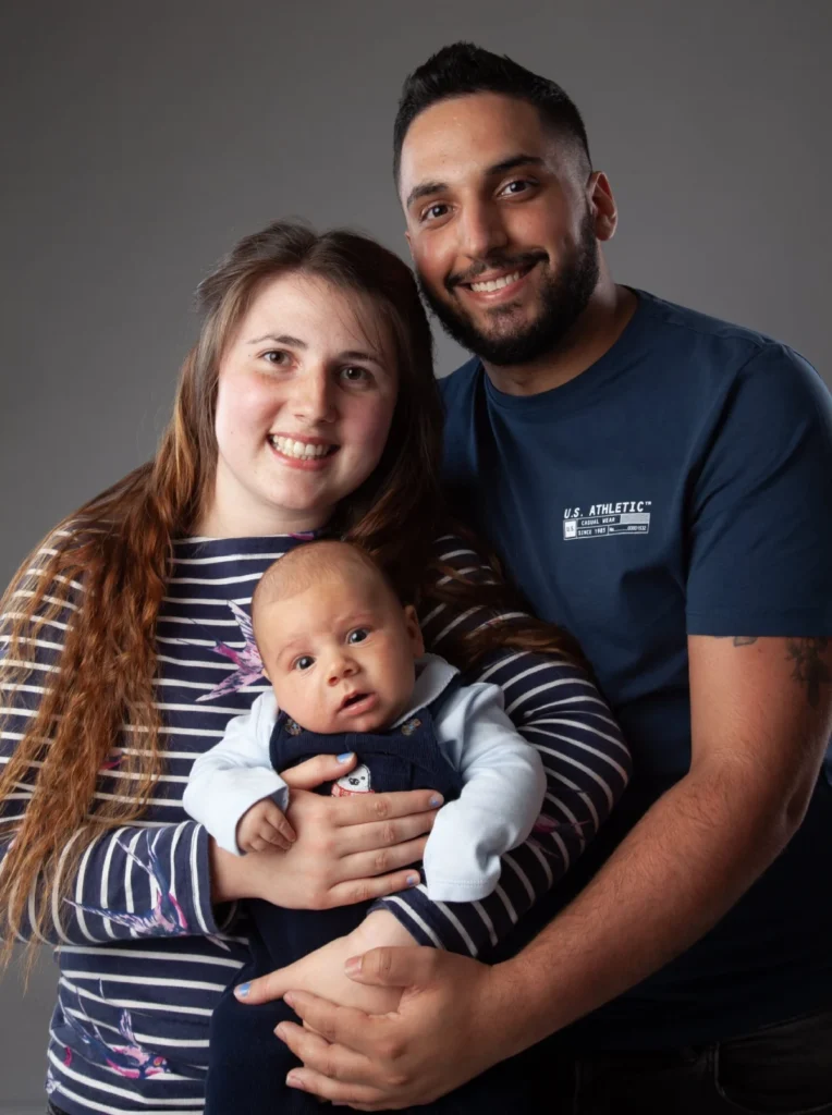 A woman and man stand close together smiling, holding a baby in front of them. The background is plain grey.